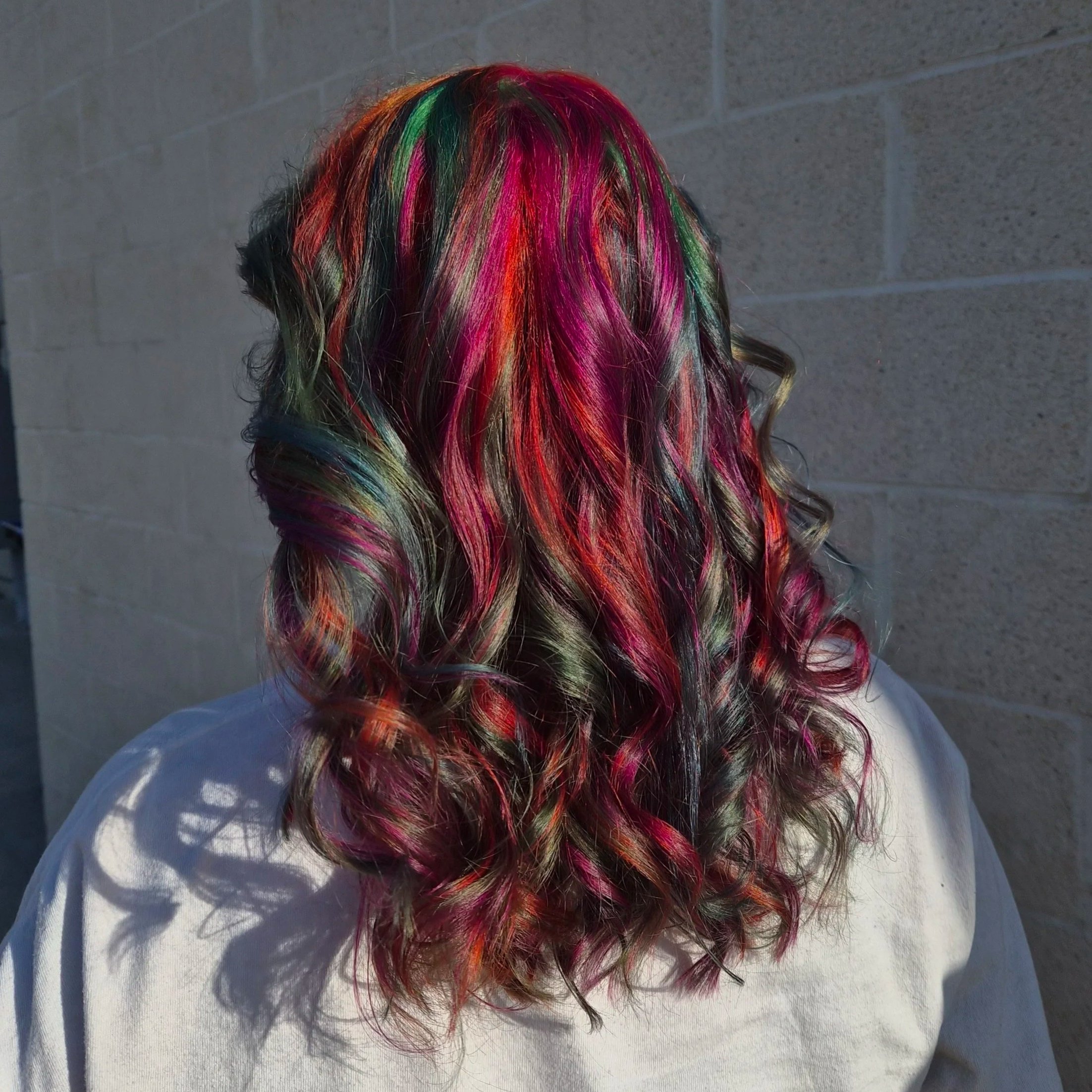 Person with long hair dyed in multiple vibrant colors including pink, red, green, and orange, standing against a brick wall.