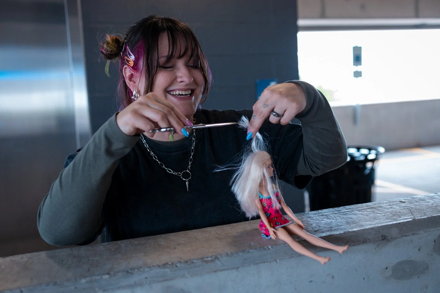 A young woman with colorful hair and jewelry smiling as she cuts hair on a Barbie doll.