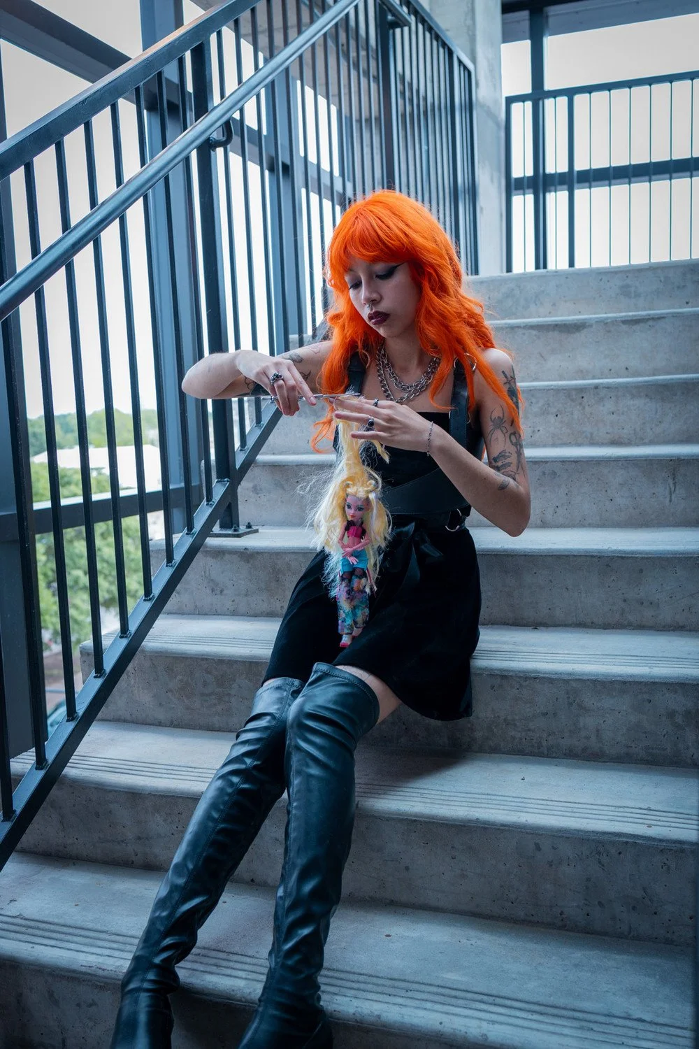 A woman with bright orange hair, tattoos, wearing a black dress, and over-the-knee black leather boots, sitting on concrete stairs. She is holding a doll with blonde hair and colorful clothing.