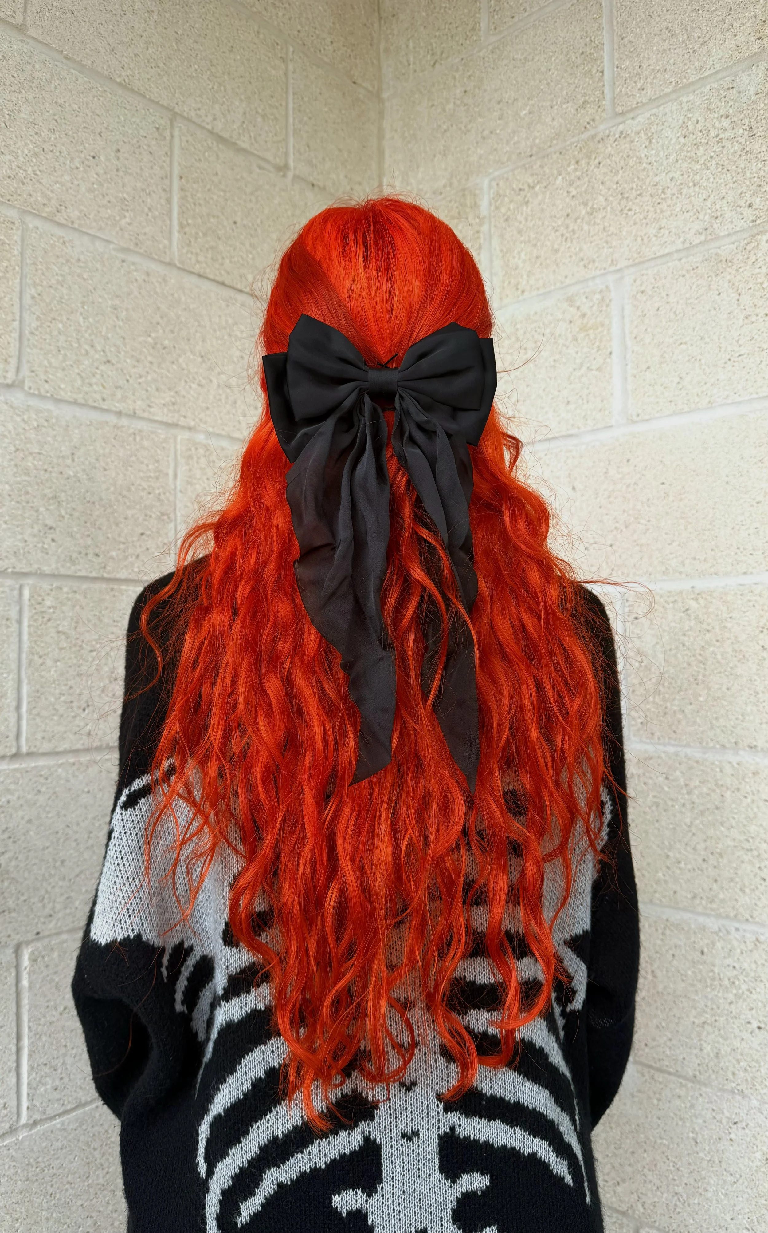 Person with long, wavy bright red hair wearing a large black bow in their hair, standing against a beige brick wall.