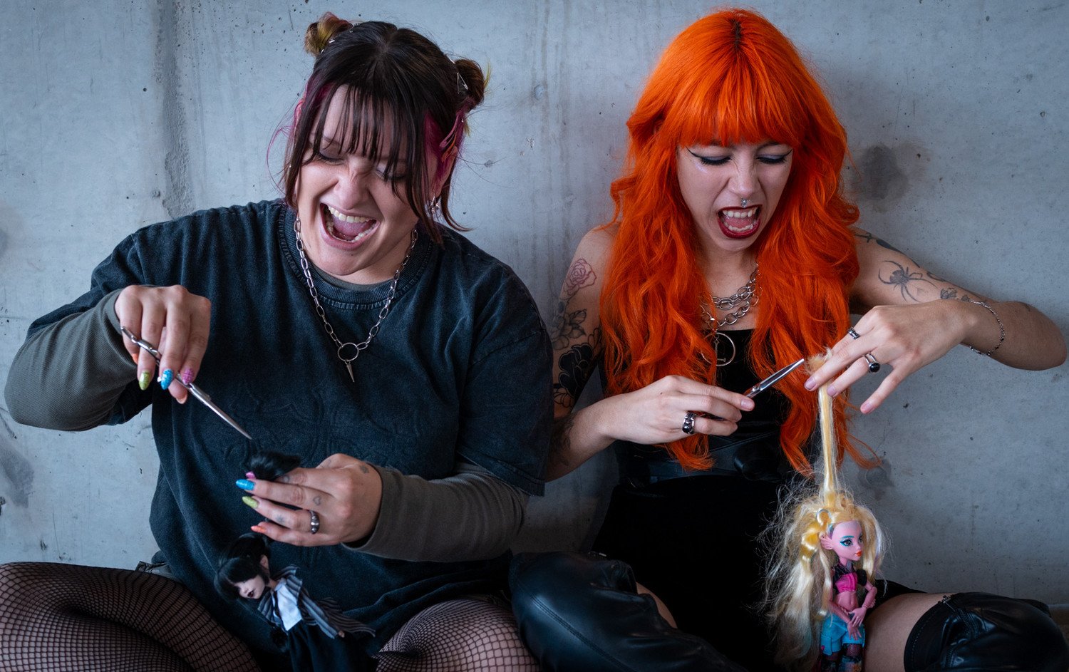 Two women with studded fishnet stockings, colorful hair, and tattoos, using scissors to cut a doll's hair with a long strand of blonde hair connecting to the doll. They are sitting against a concrete wall, laughing and styling the doll.