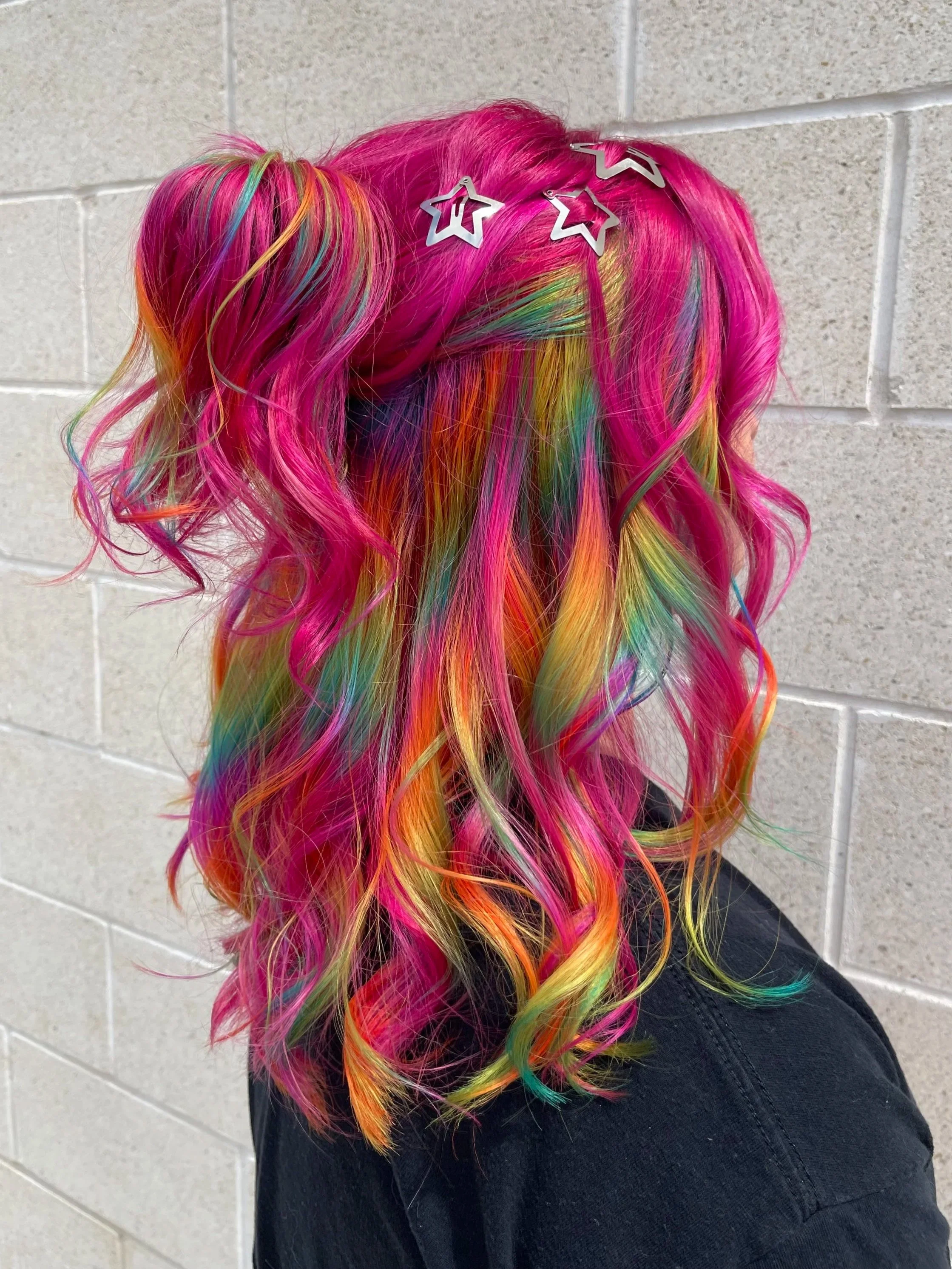 Colorful rainbow curly hair decorated with star-shaped hair clips, against a beige tiled wall.