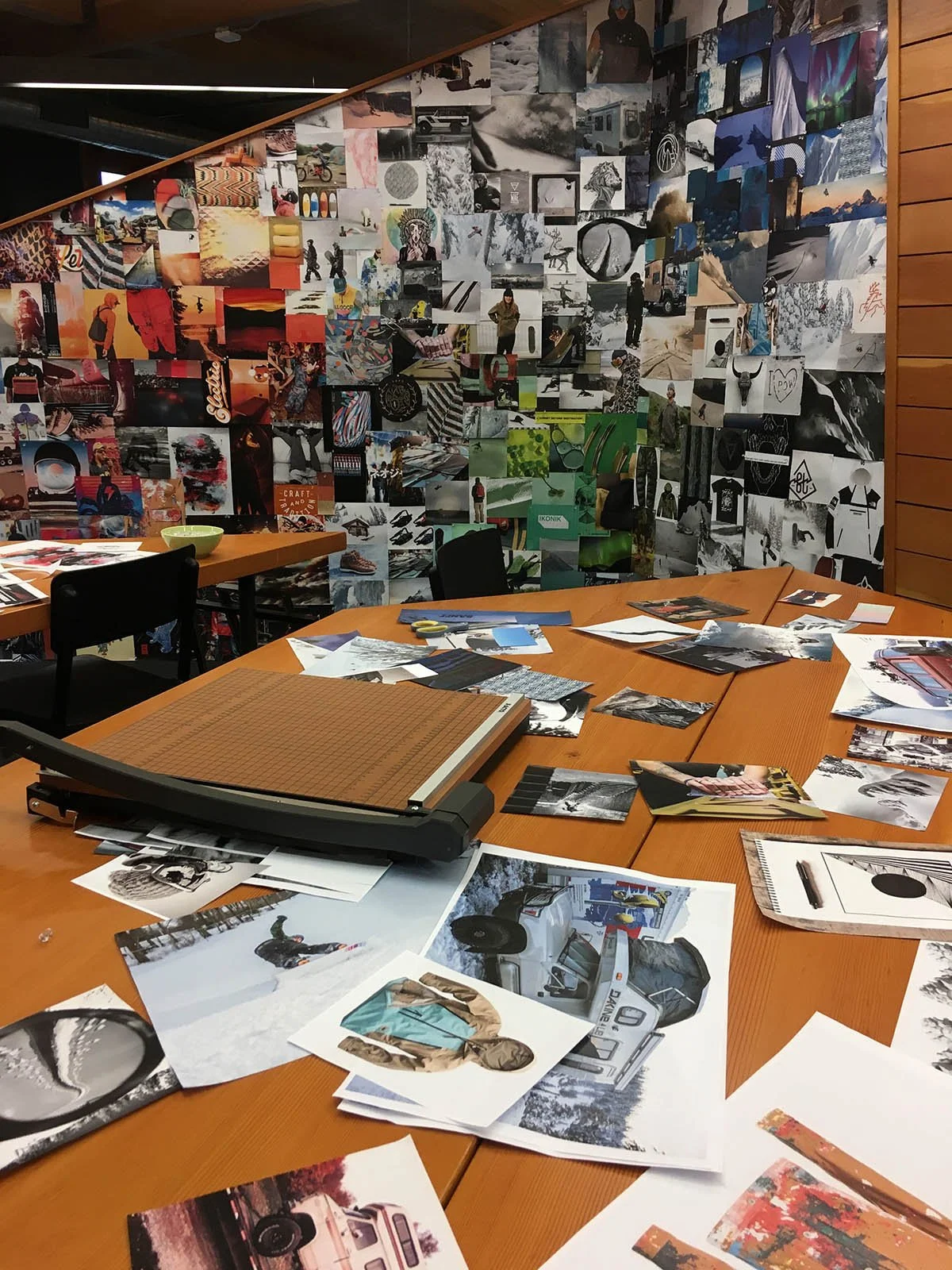 A room with a large wall covered in a collage of various photographs and images. There are tables cluttered with pictures, photos, and art supplies, indicating an art or creative workspace.