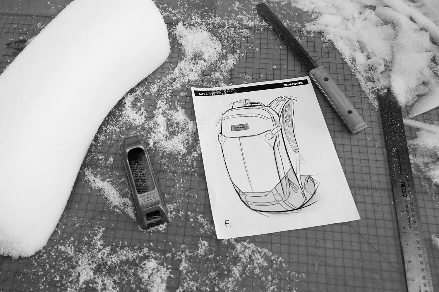 Engineering workspace with a technical drawing of a backpack, a utility knife, a ruler, and foam material, scattered on a cutting mat.