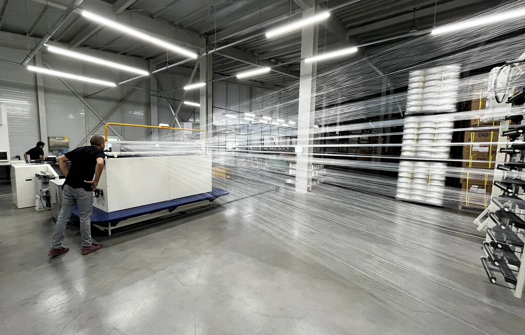 People operating a machine in a warehouse, with moving strings of plastic or film being produced or processed.
