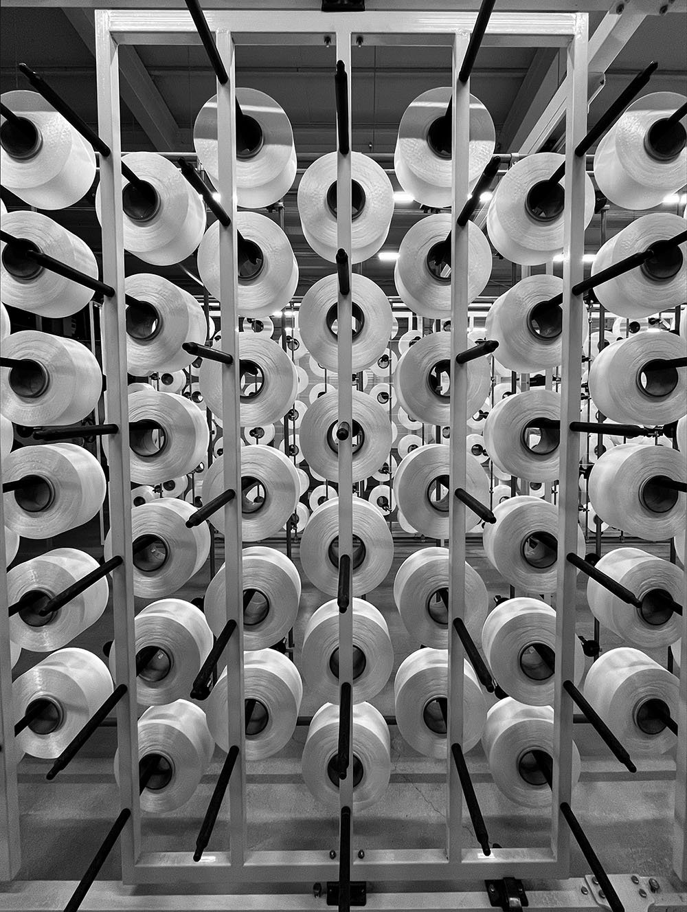 A display of multiple large rolls of fabric or yarn arranged on black rods within a white metal rack, in a store or factory setting.