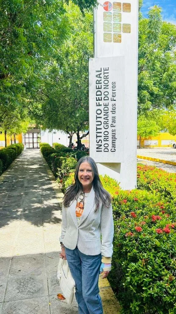 A woman with long dark hair wearing a striped blazer, orange patterned shirt, and blue jeans standing in front of a sign at the Instituto Gran Departamento campus of the Federal University of Pernambuco, with green trees and bushes in the background.
