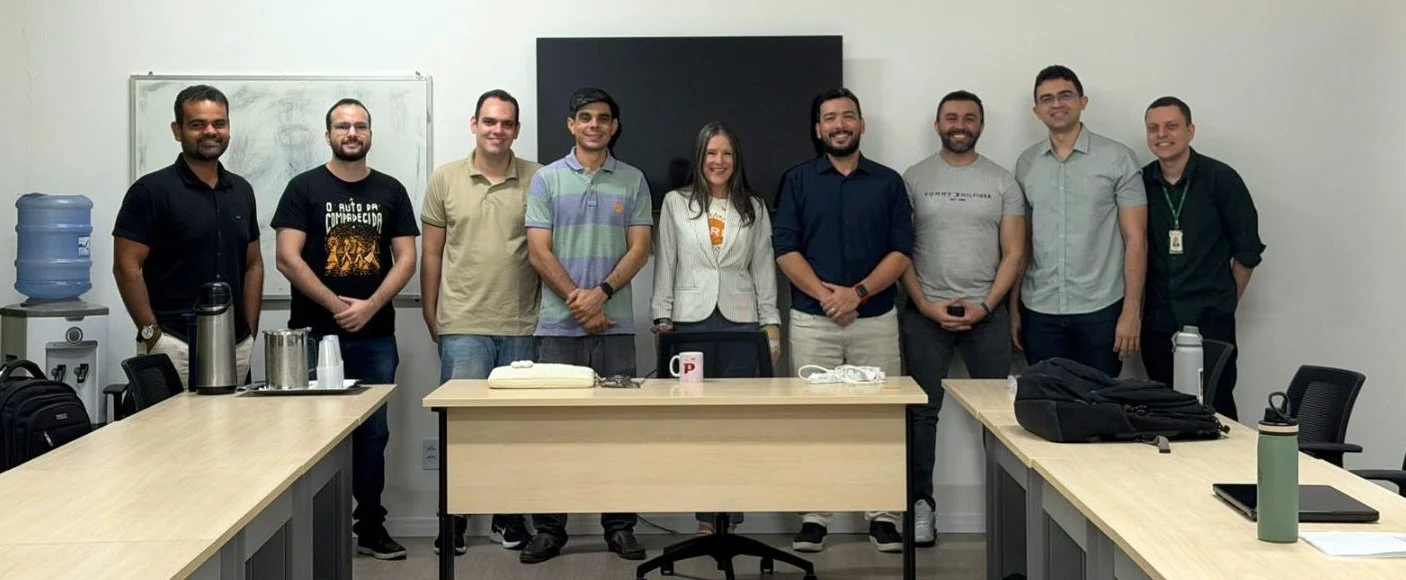 Group of nine people standing in a meeting room behind a long conference table with coffee mugs, water jug, backpacks, and laptops, smiling at the camera.