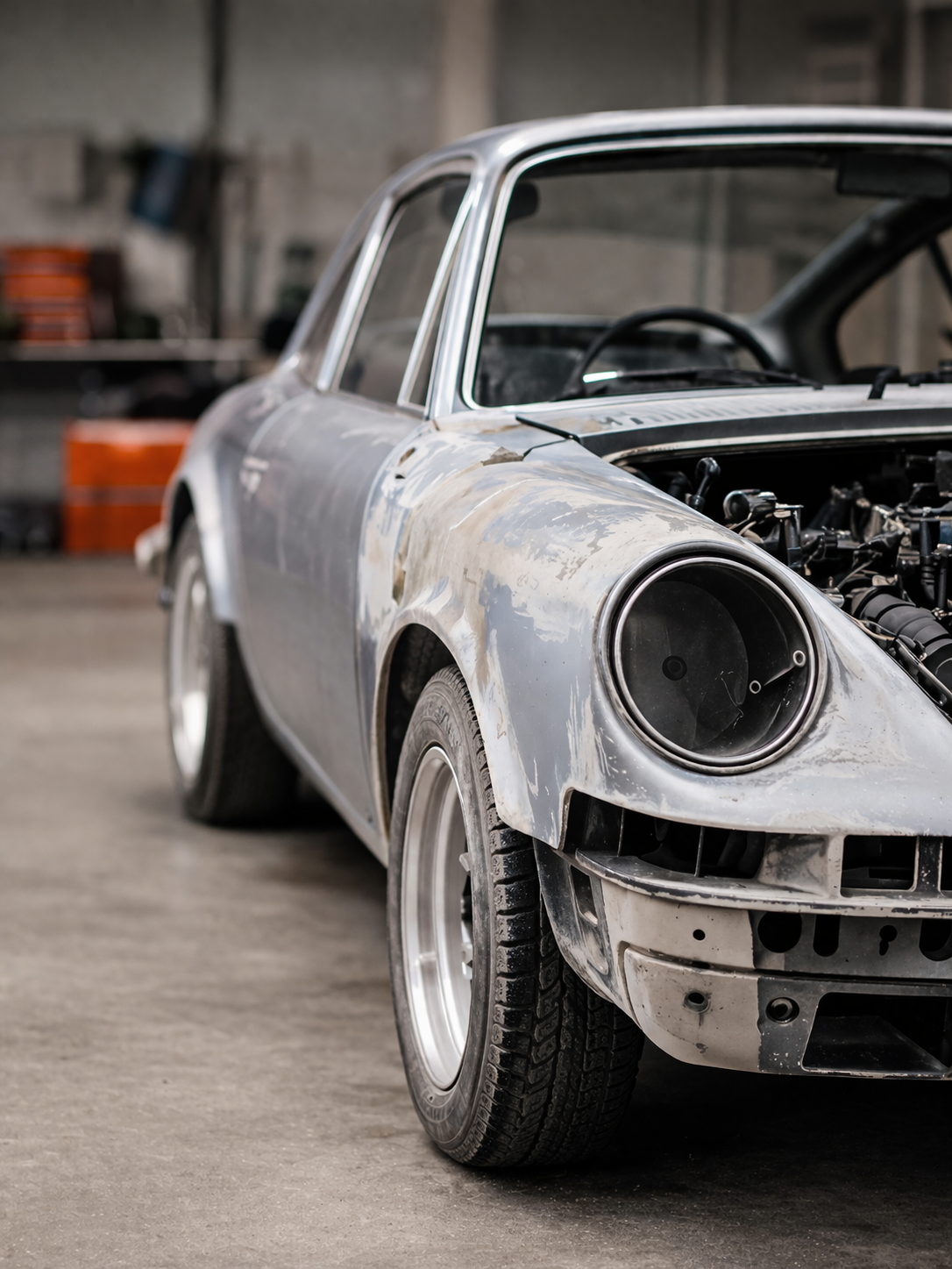 Een oude, onafgewerkte sportwagen in een garage, zonder koplamp en met zichtbaar motorcompartiment.