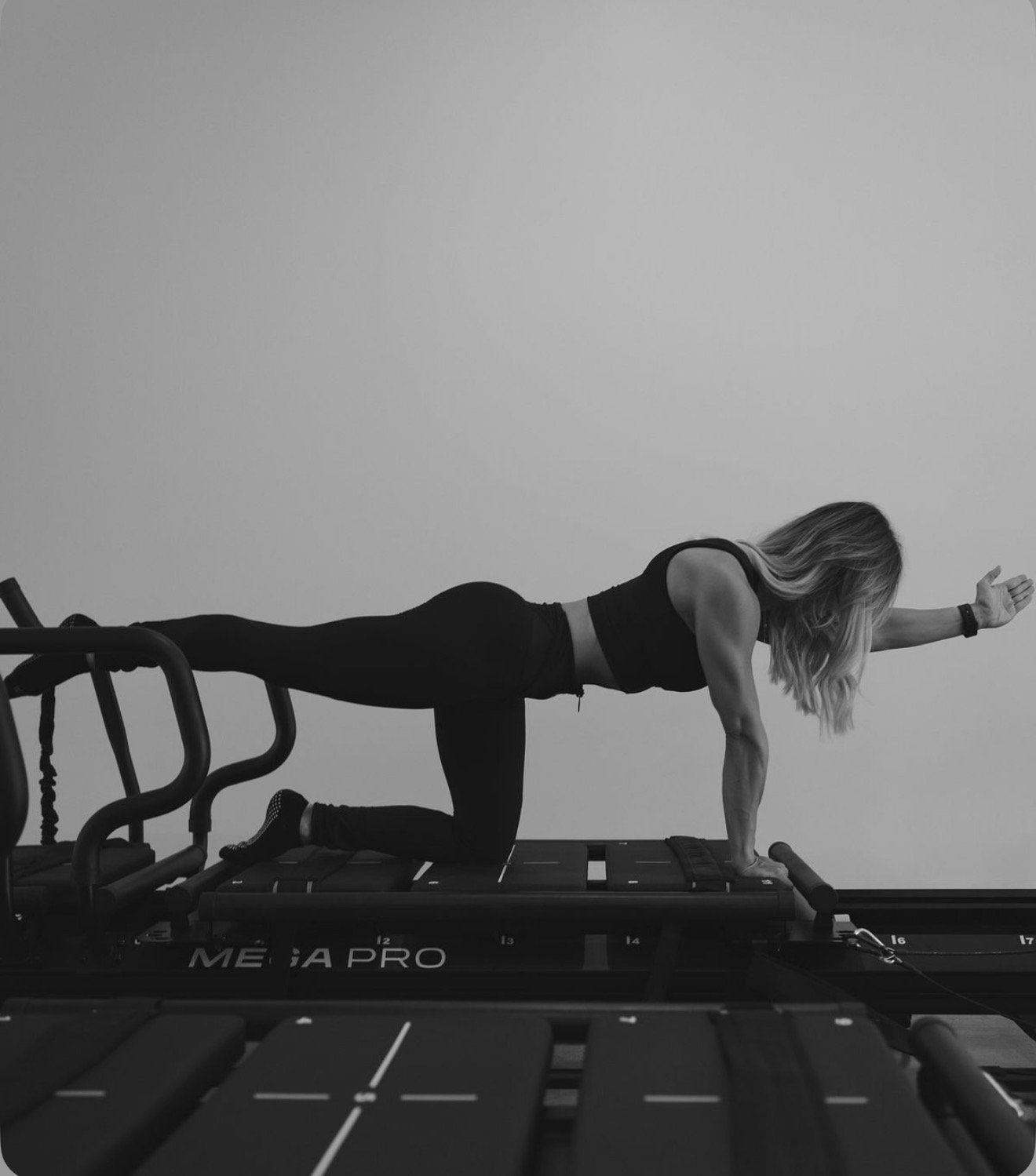 A woman doing lagree on a reformer, reaching forward with one arm while balancing on one knee, in a minimalist studio with plain walls.