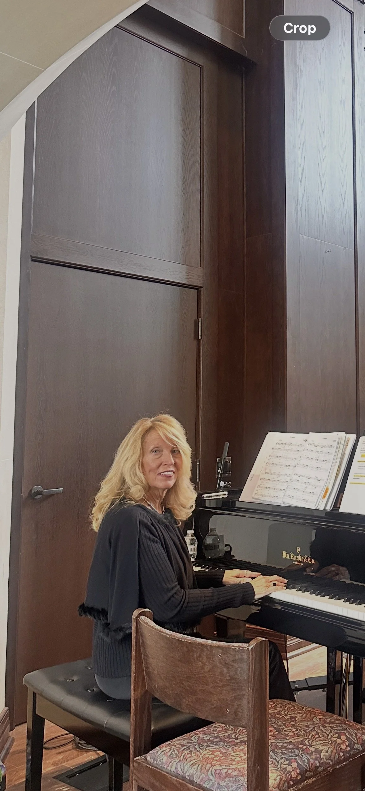 A woman with blonde hair is sitting at a black grand piano in a room with dark wooden walls, looking towards the camera. There are sheet music and a music stand on the piano.