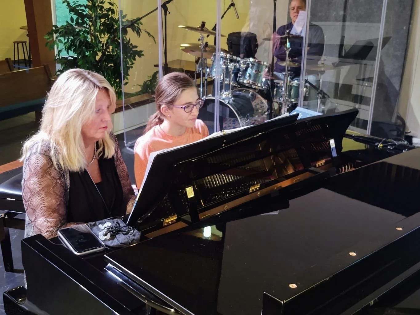 Two women sit at a grand piano, playing music together, with a drummer in a soundproof room behind a glass wall in the background.