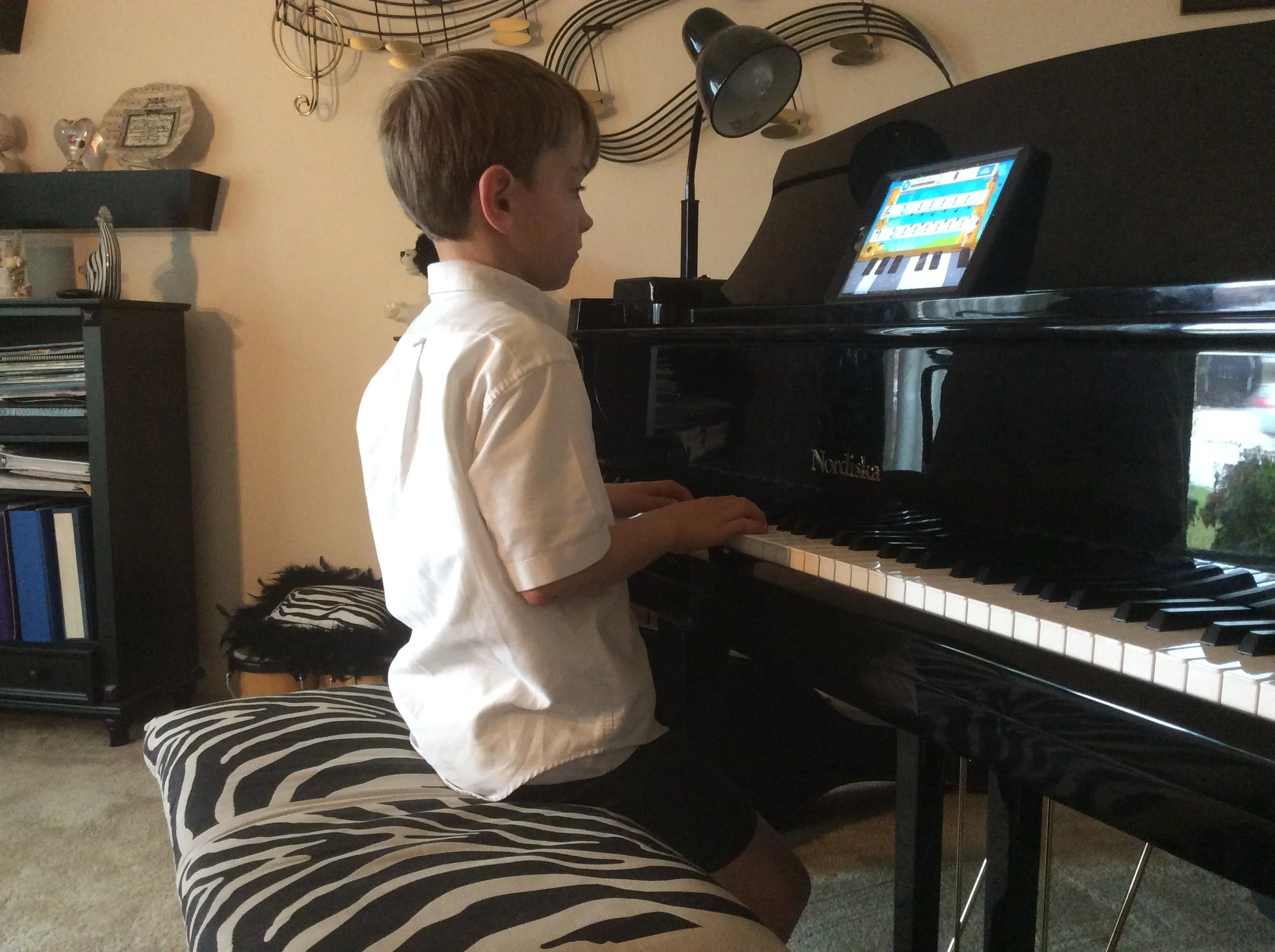 A young boy playing a black grand piano in a living room, using a tablet on the music stand. The room has a zebra-striped bench, a bookshelf, and decorative shelves on the wall.
