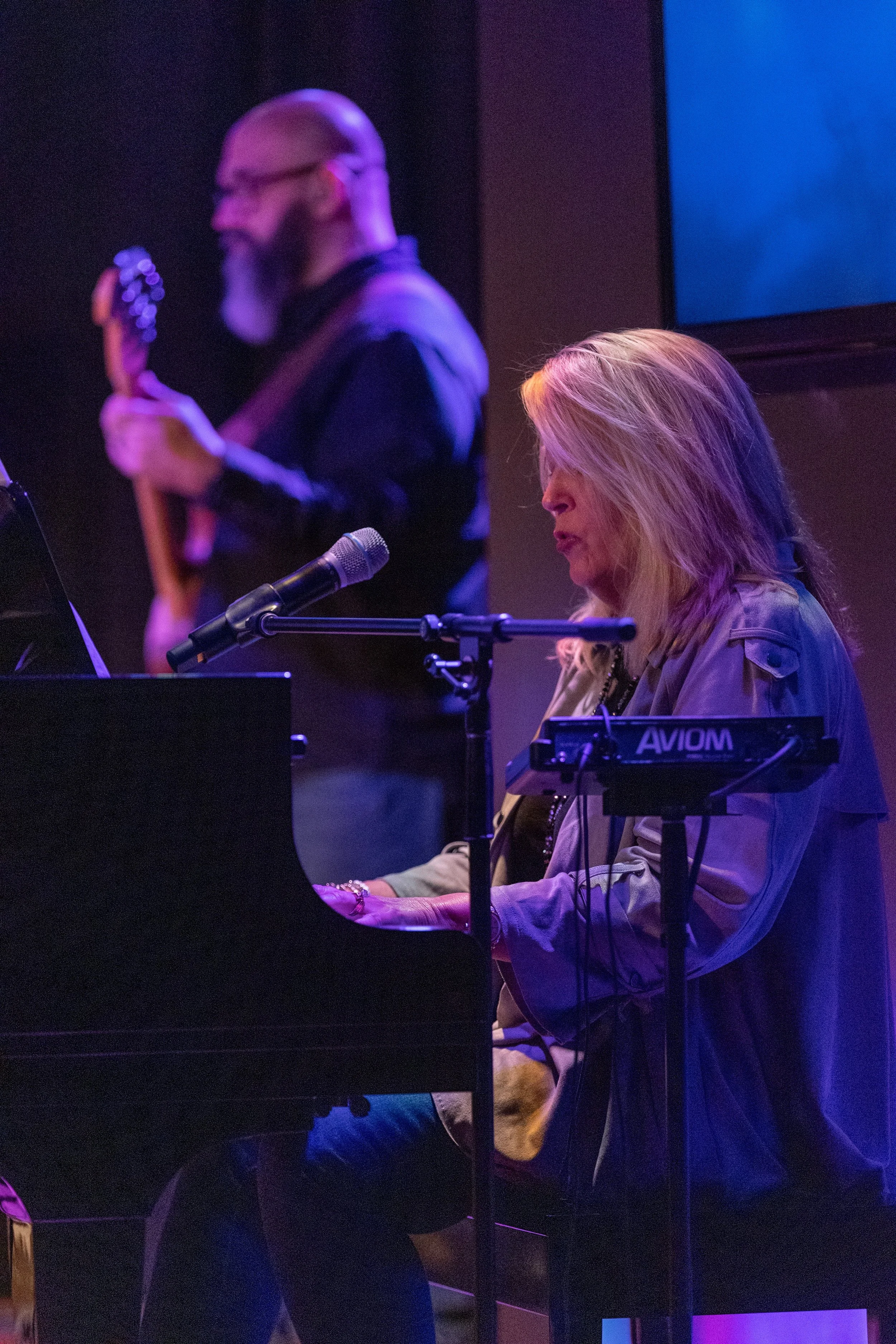 A woman playing the piano and a man playing the guitar during a live performance, with stage lighting.