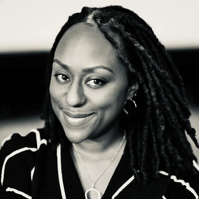 A smiling woman with long dark locs, a nose piercing, and a necklace, in a black and white portrait.