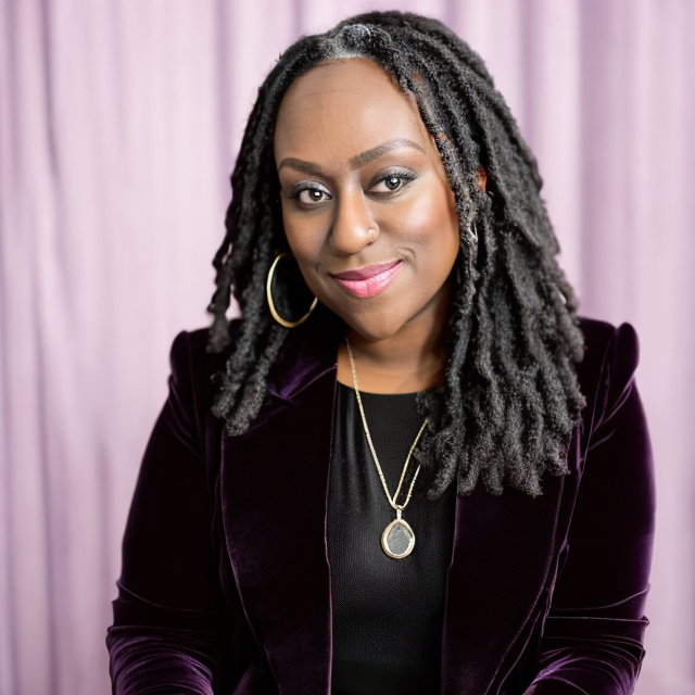 A woman with long, dark locs and hoop earrings, wearing a dark velvet blazer and a black top, standing in front of a pink curtain, smiling at the camera.