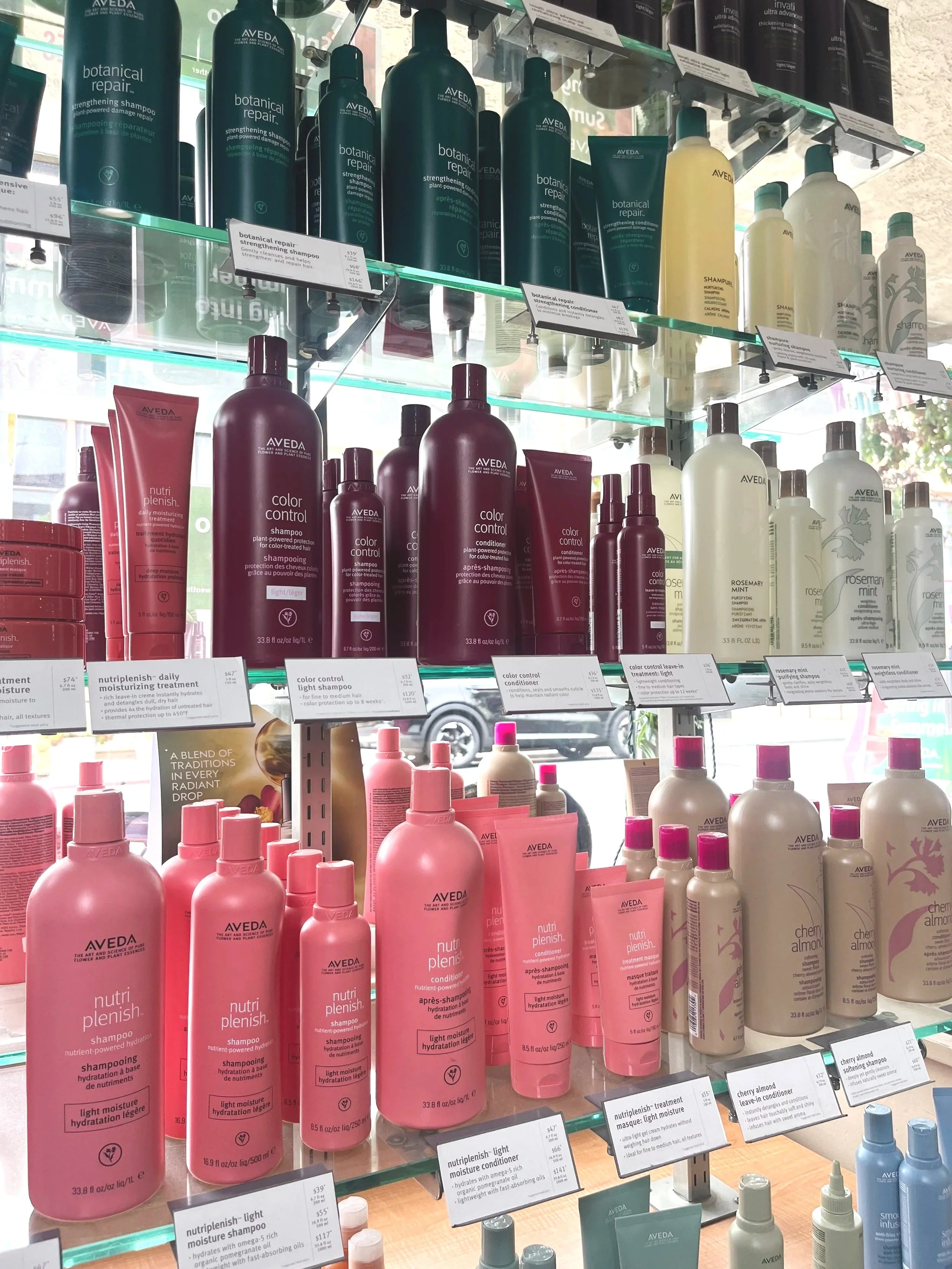 Display of Aveda hair care products on shelves, including shampoos, conditioners, and treatments in various colors such as pink, white, teal, and black, with product labels.