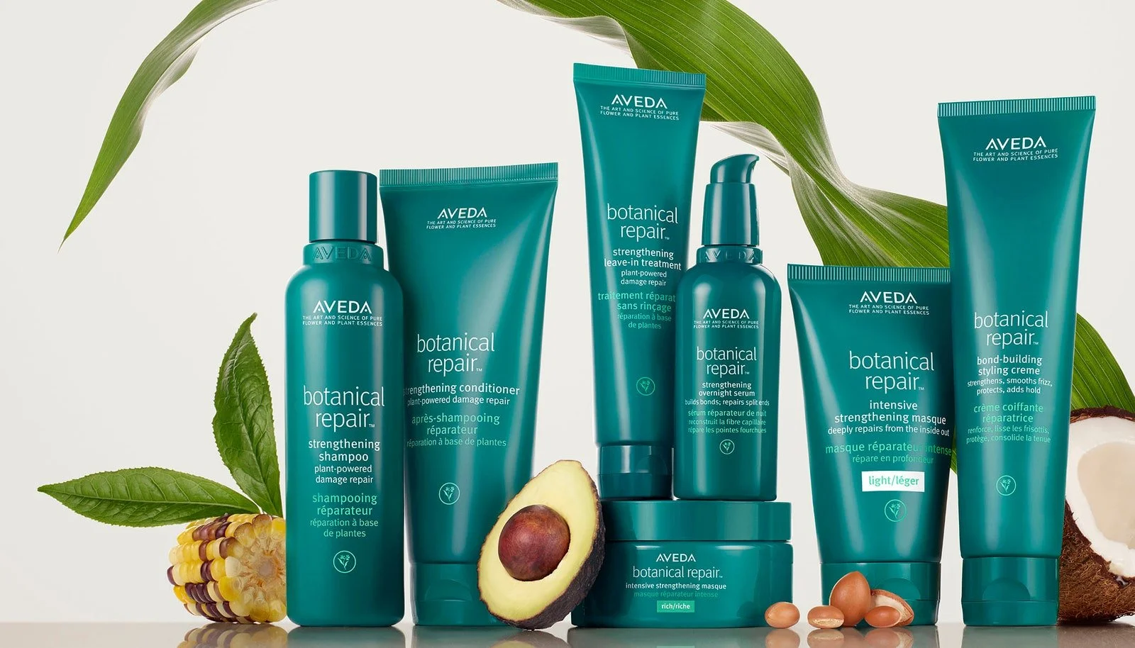 A collection of Aveda Botanical Repair hair care products, including shampoo, conditioner, leave-in treatment, strengthening serum, mask, and styling crème, arranged with tropical fruits and green leaves.