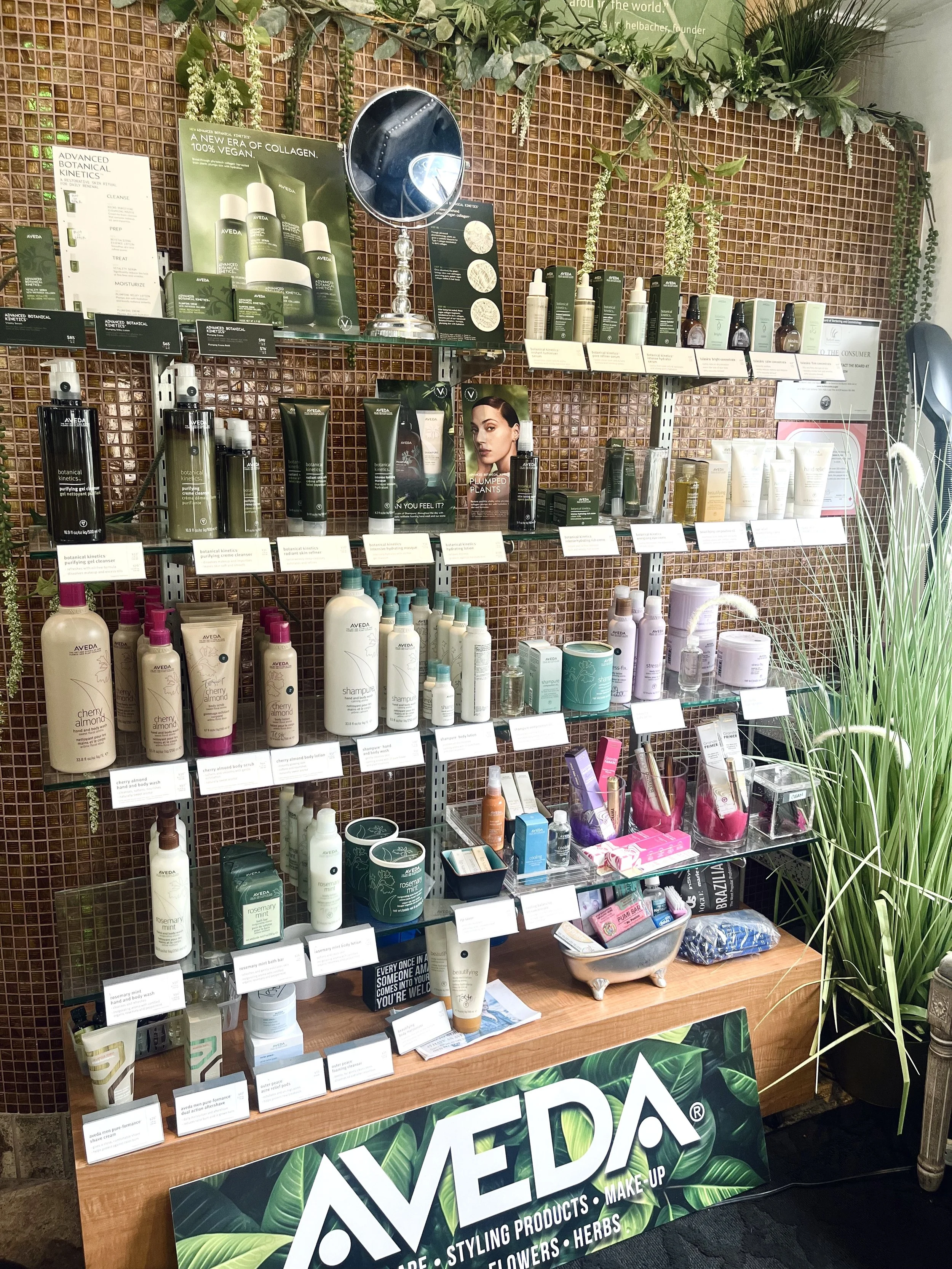Display of AVEDA skincare and haircare products at a store or event, with shelves filled with bottles, jars, and boxes, partially decorated with green plants, against a textured brown tile wall.