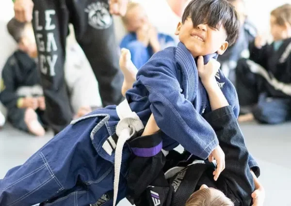 Children participating in a Brazilian Jiu-Jitsu class, with one child in a blue gi demonstrating a grappling move on another child in a black gi, in a martial arts training facility.