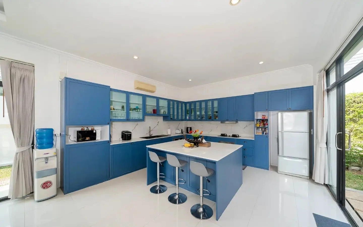 Modern kitchen with blue cabinets, white countertops, and a kitchen island with three gray stools, adjacent to sliding glass doors. Villa Lotus Heaven, Cherng Talay, Phuket
