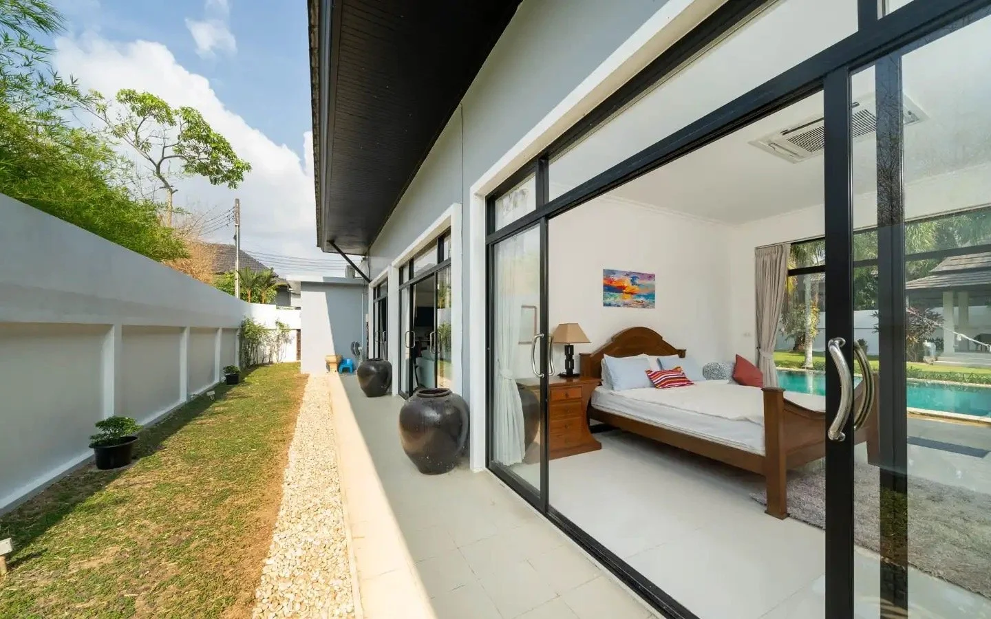 View of a modern house with a glass sliding door, showing a bedroom with a wooden bed, a nightstand with a lamp, and a colorful painting on the wall. Outside, there is a backyard with a swimming pool, green lawn, and some trees. Villa Lotus Heaven, C