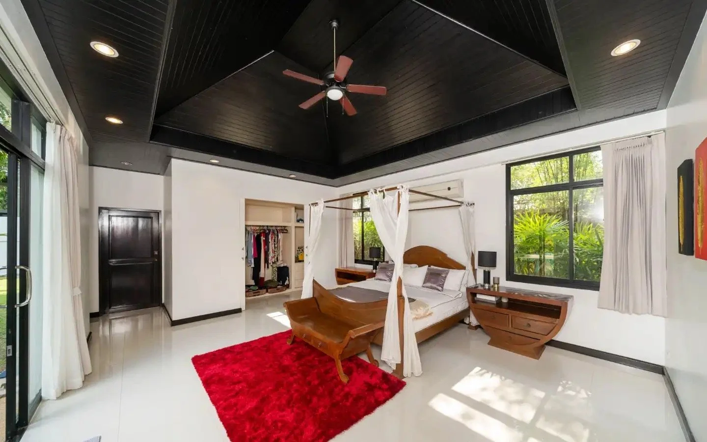 A brightly lit bedroom with a wooden four-poster bed, dark wood nightstands, a red area rug, and windows showing greenery outside. Villa Lotus Heaven, Cherng Talay, Phuket