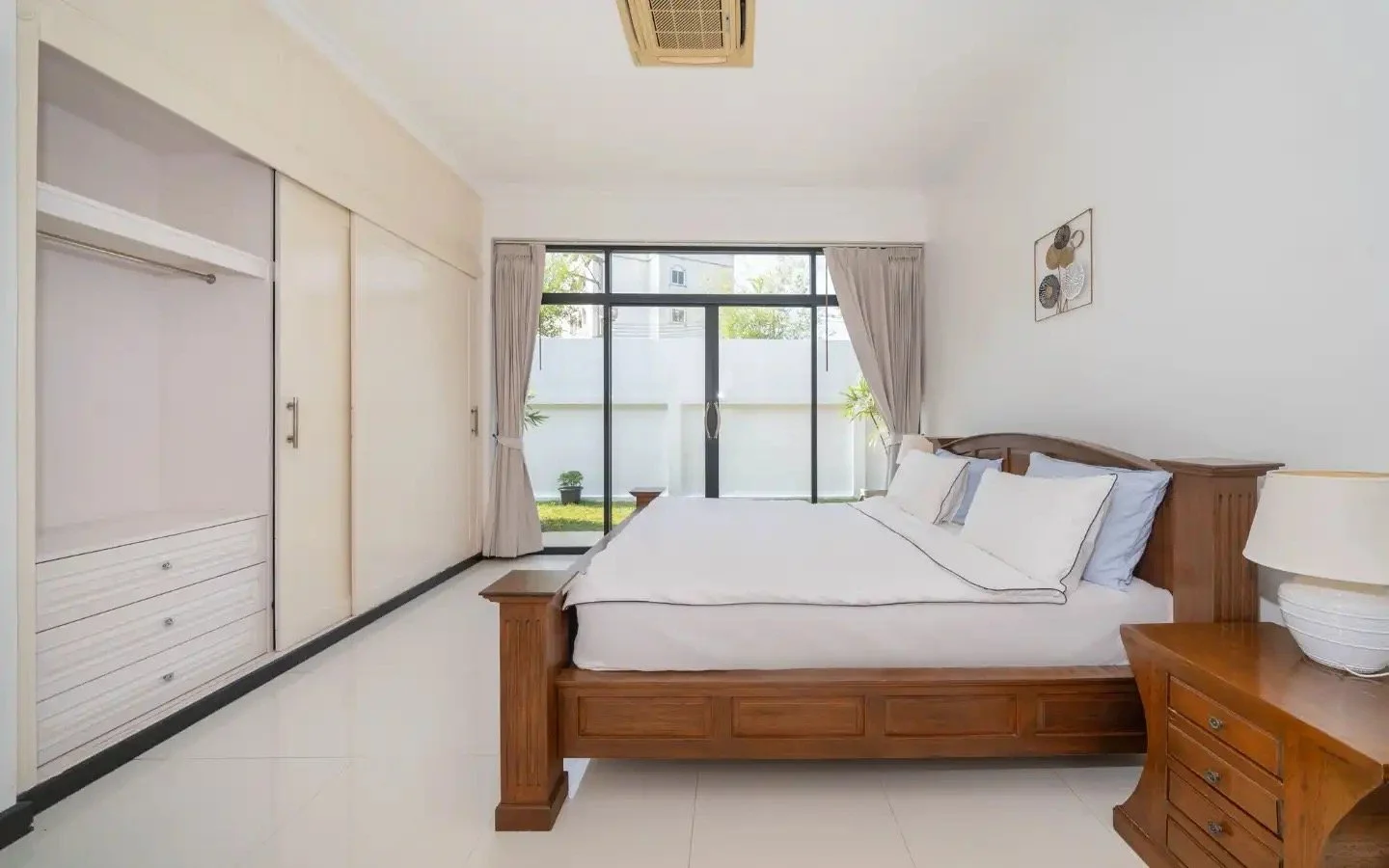 A bedroom with a wooden bed frame, white bedding, and pillows; a nightstand with a lamp; a large glass door with beige curtains leading to an outdoor garden; and a built-in closet on the left side. Villa Lotus Heaven, Cherng Talay, Phuket