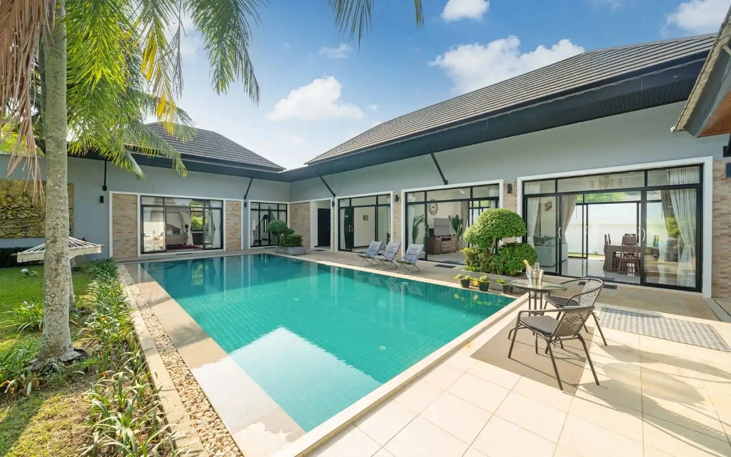 Modern house with a swimming pool surrounded by patio chairs and lush green plants under a partly cloudy sky. Villa Lotus Heaven, Cherng Talay, Phuket