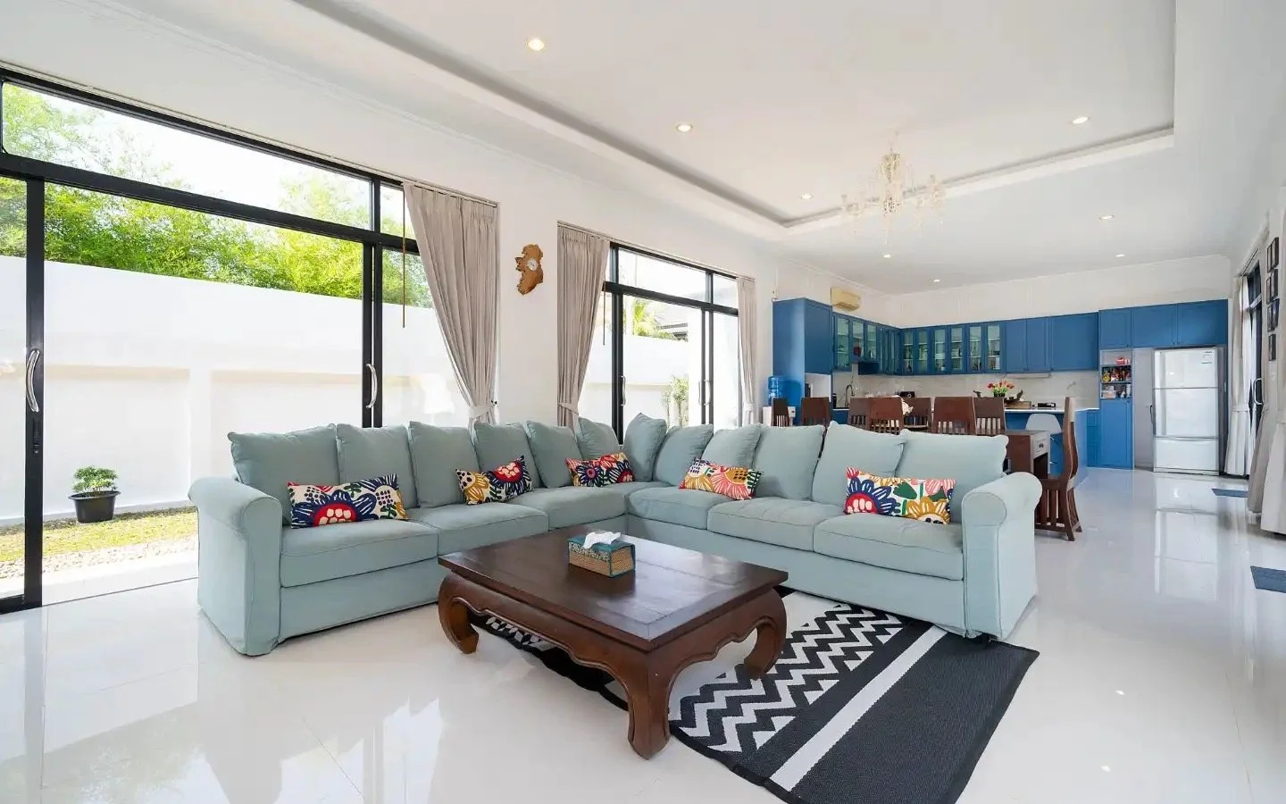 Living room with large windows, light blue sectional sofa with colorful patterned pillows, wooden coffee table, black-and-white patterned rug, open kitchen with blue cabinets, white appliances, and dining table.