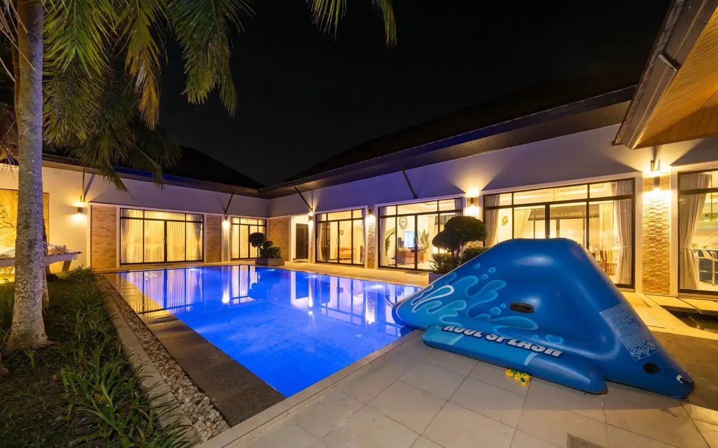 A backyard at night with a lit swimming pool, a blue inflatable water slide, tall palm trees, and a modern house with large glass sliding doors and warm interior lighting. Villa Lotus Heaven, Cherng Talay, Phuket