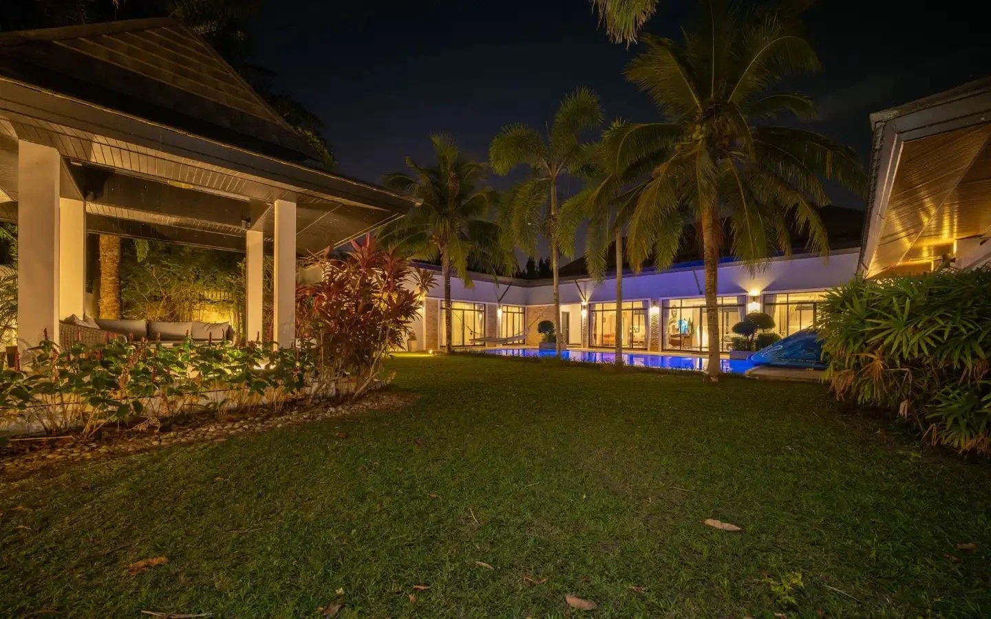 Night view of a luxurious house with a backyard, tall palm trees, well-lit with warm lighting, and a swimming pool reflecting lights, surrounded by lush greenery. Villa Lotus Heaven, Cherng Talay, Phuket