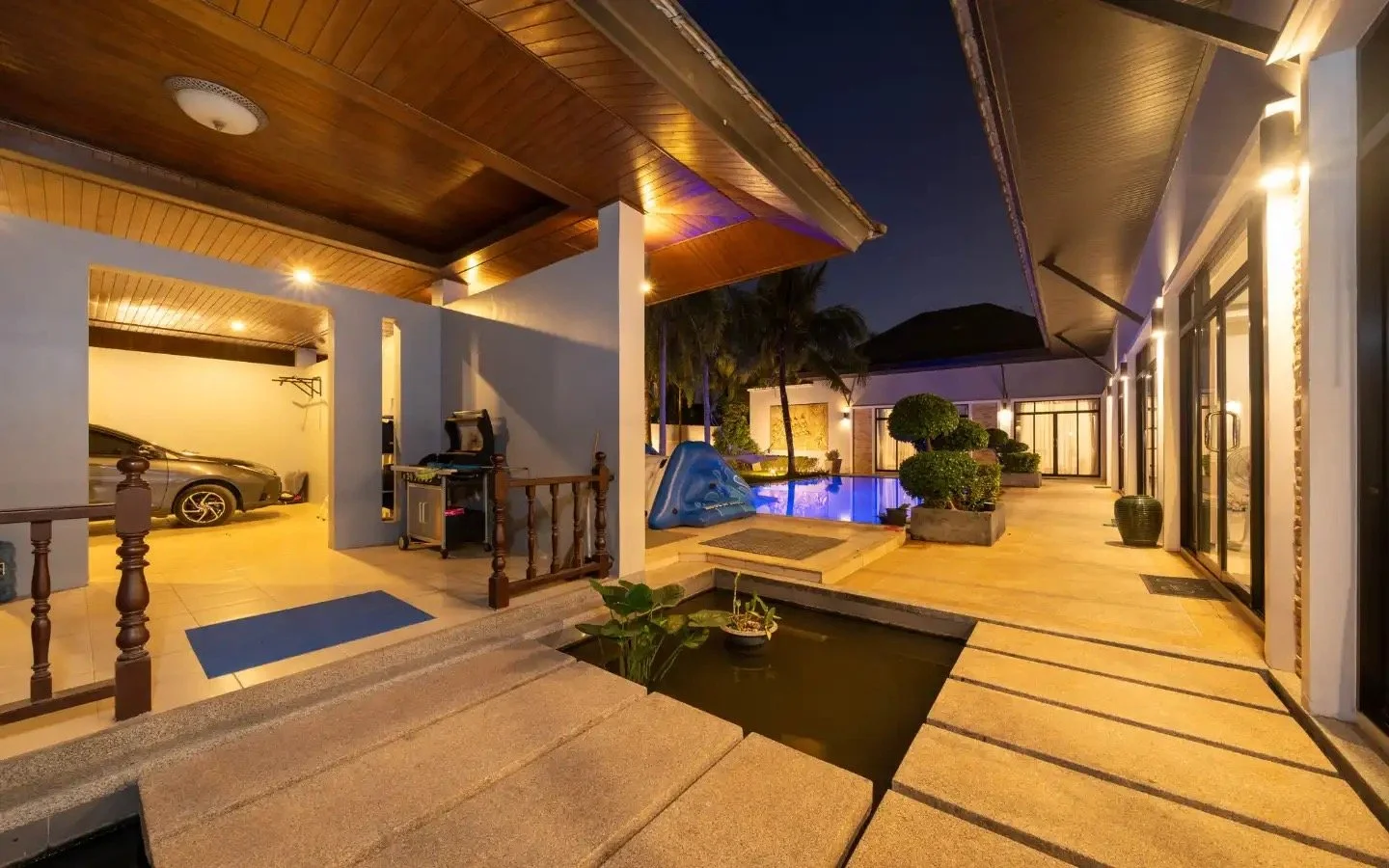 Night view of a modern house's backyard with a swimming pool, potted plants, a children's slide, and a car parked in a garage. Villa Lotus Heaven, Cherng Talay, Phuket