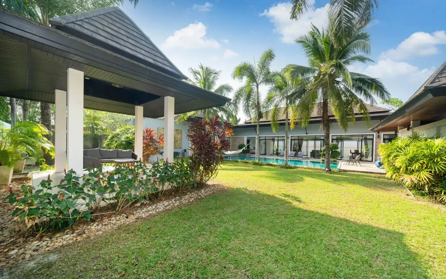 A tropical backyard with a lush lawn, palm trees, and a modern house featuring large glass sliding doors and a pool.