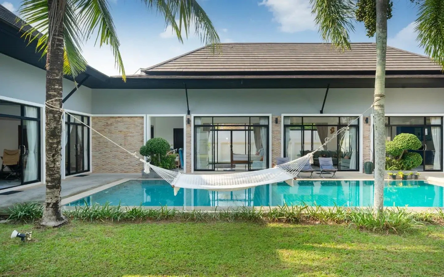 A modern house with large glass sliding doors, a swimming pool in front, a white hammock tied between two palm trees, and a well-manicured lawn in a tropical setting. Villa Lotus Heaven, Cherng Talay, Phuket