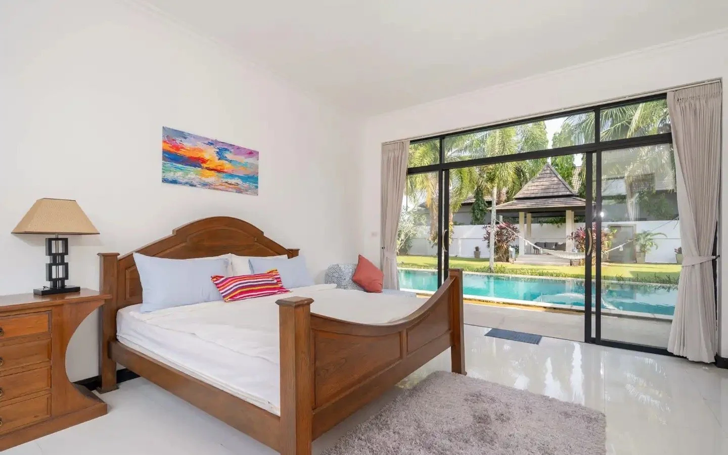 Bedroom with wooden bed, colorful striped pillow, white bedding, bedside table with lamp, artwork above the bed, large sliding glass door opening to pool and tropical garden. Villa Lotus Heaven, Cherng Talay, Phuket