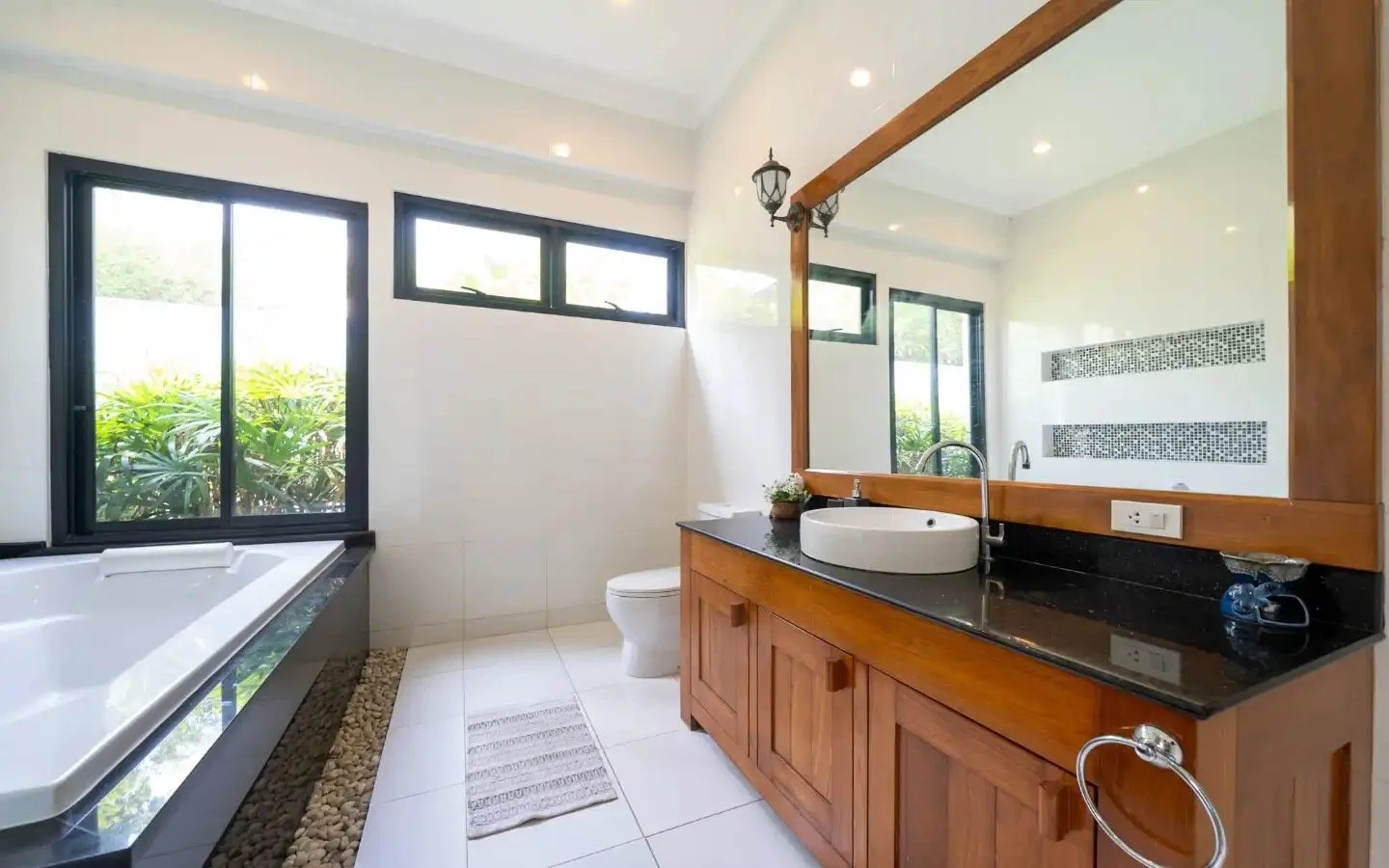 A modern bathroom with a large bathtub, black framed windows, a wooden vanity with a black countertop, a white vessel sink, a large mirror, and a toilet, illuminated by natural light. Villa Lotus Heaven, Cherng Talay, Phuket