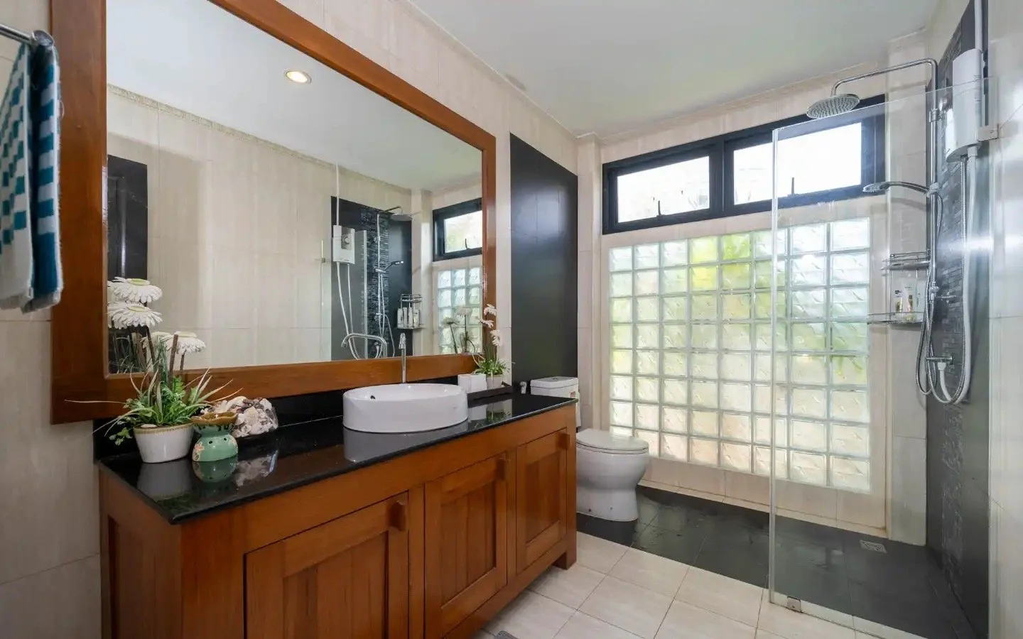 Bathroom with wooden vanity, large mirror, glass-block window, shower area with rain showerhead, toilet, and decorative plants. Villa Lotus Heaven, Cherng Talay, Phuket