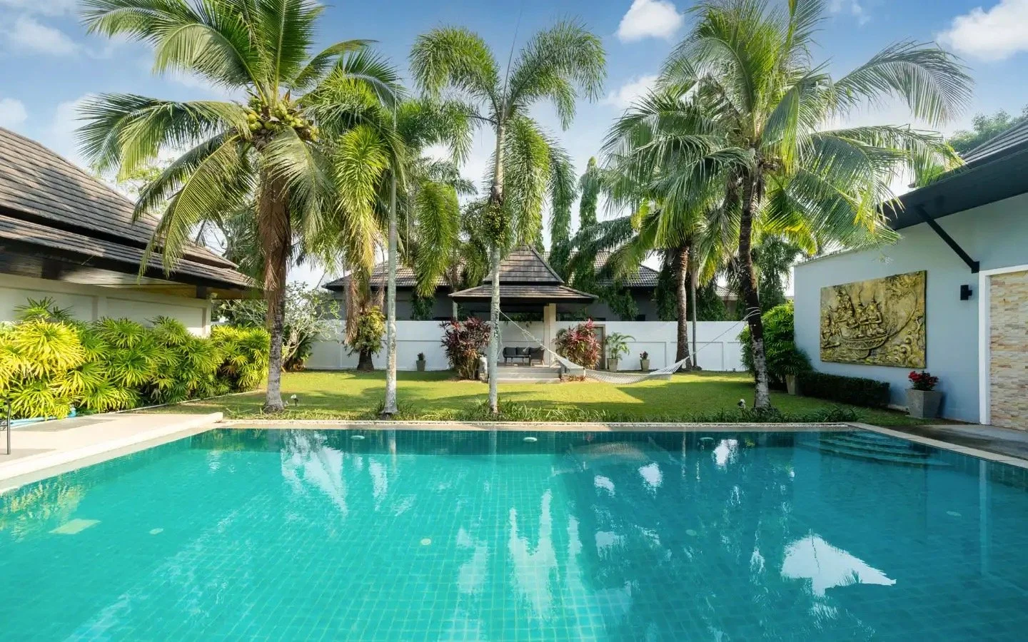 A backyard with a swimming pool, surrounded by palm trees, a grassy area, and a small pavilion with outdoor seating, all enclosed by white fencing. Villa Lotus Heaven, Cherng Talay, Phuket