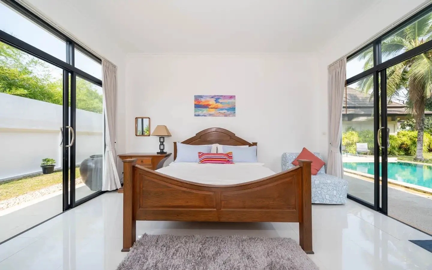 Bedroom with large windows showing a backyard with pool and tropical plants, wooden bed with white bedding, side table with lamp, and a patterned armchair. Villa Lotus Heaven, Cherng Talay, Phuket