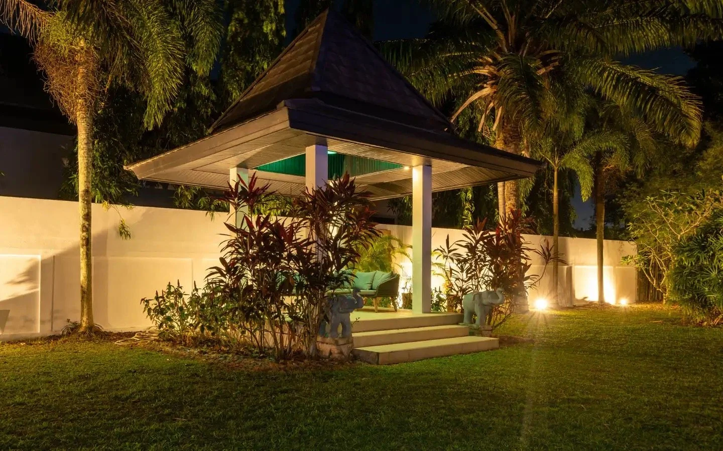A nighttime outdoor scene of a small, open-sided pavilion with a pointed roof, surrounded by lush tropical plants and trees, illuminated by landscape lighting, with steps leading up to it and two small elephant statues on either side of the stairs. V