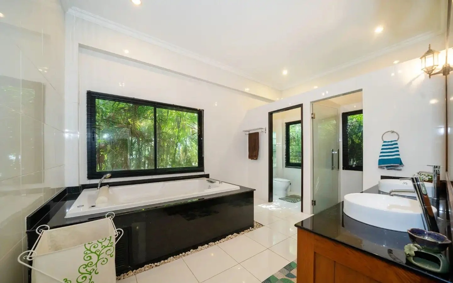 Bright bathroom with a large window showing greenery outside, a black bathtub, a toilet in a separate room, a white sink on a wooden cabinet, and a towel hanging on the wall. Villa Lotus Heaven, Cherng Talay, Phuket