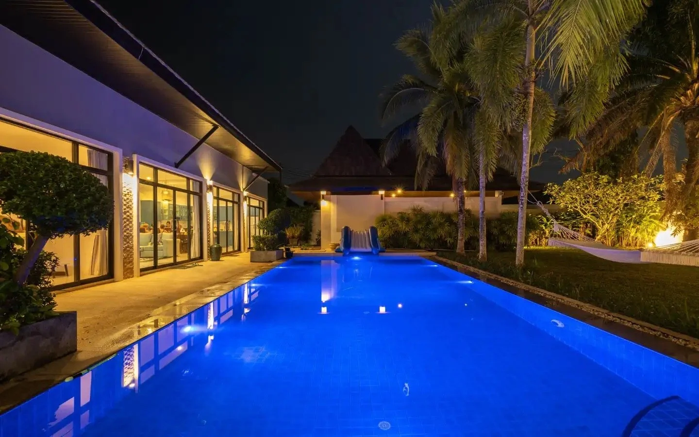 Night view of an illuminated swimming pool surrounded by tropical trees and a house with large glass doors. Villa Lotus Heaven, Cherng Talay, Phuket