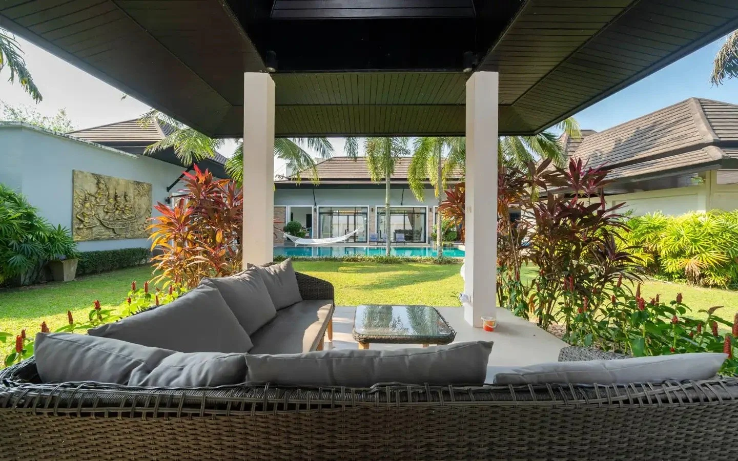 Outdoor seating area under a dark roof with white support columns, overlooking a backyard with green lawn, tropical plants, a pool with a hammock, and a house with large windows. Villa Lotus Heaven, Cherng Talay, Phuket