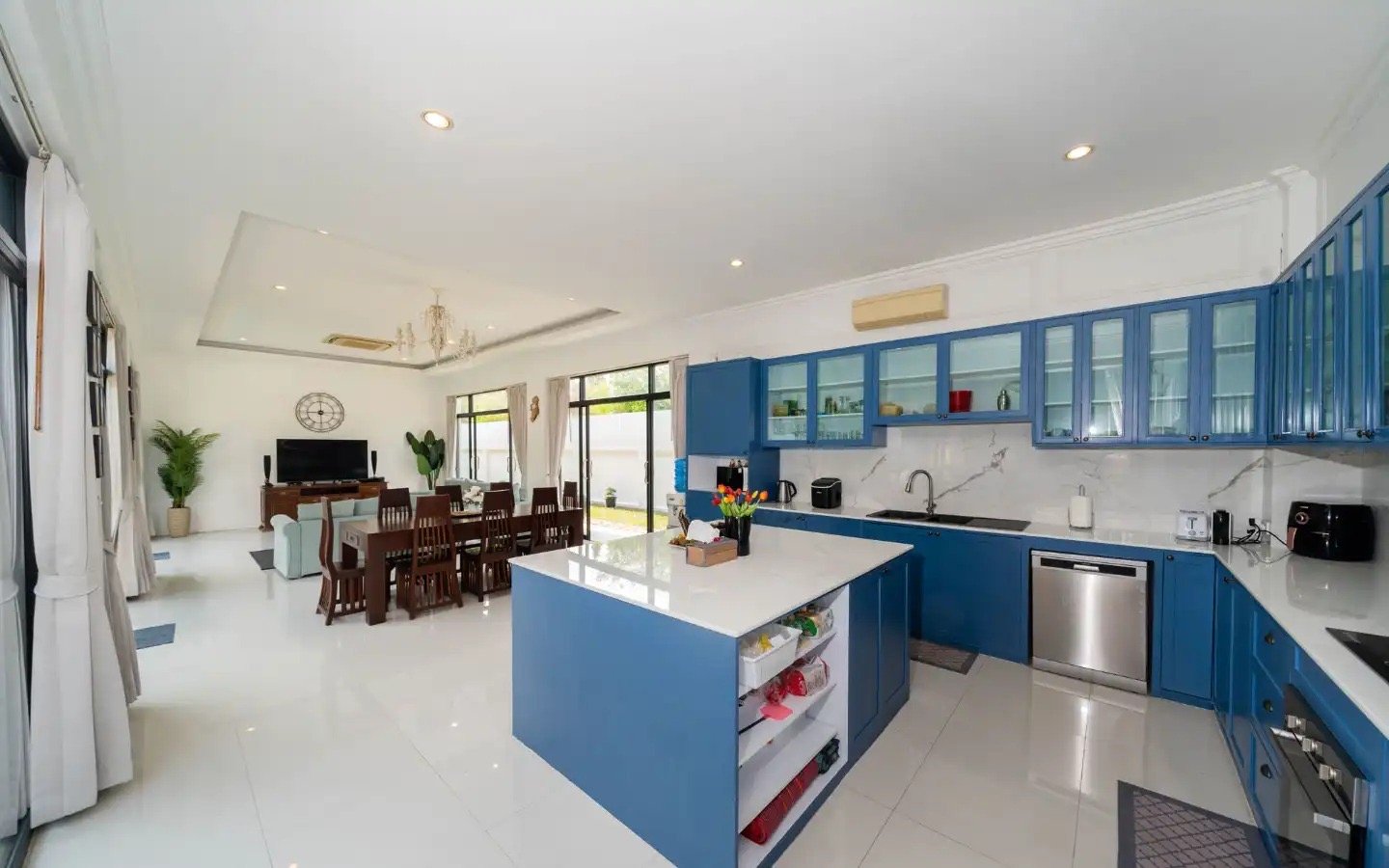 Open-concept living and kitchen area with white walls, blue cabinets, white countertops, and a kitchen island. The dining table has multiple wooden chairs, and the living area features a wall-mounted flat-screen TV, a sofa, and large windows that let