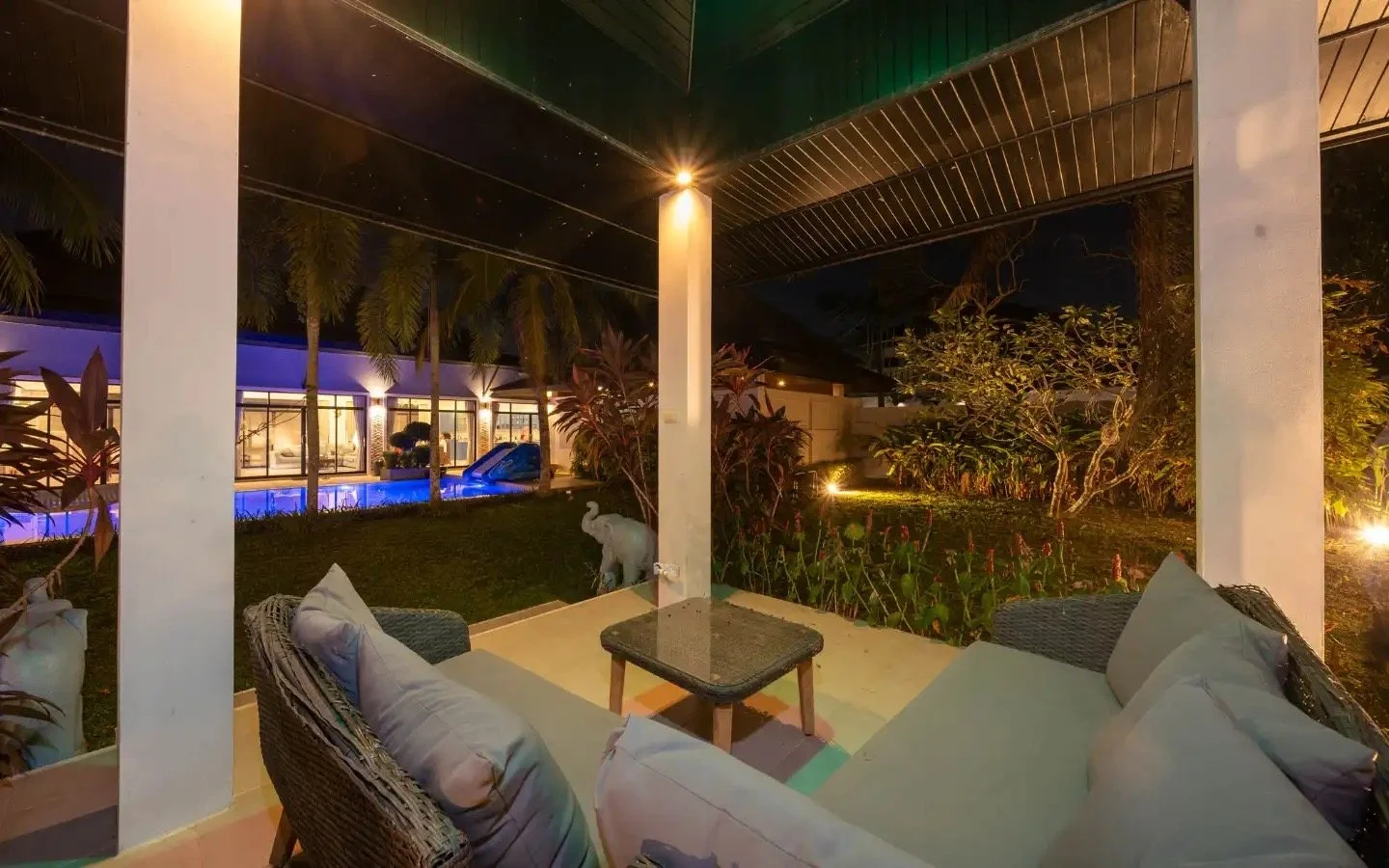 Night outdoor patio with beige cushioned wicker furniture, decorative plants, white elephant statue, and garden lighting, overlooking a pool with blue lighting and a house with large windows. Villa Lotus Heaven, Cherng Talay, Phuket