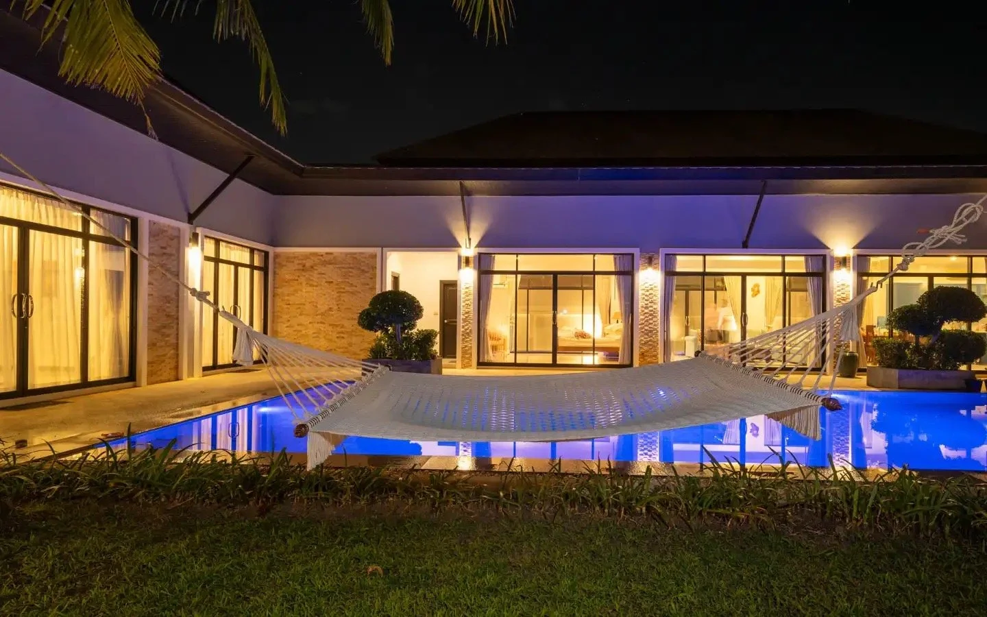 Nighttime view of a modern house with large glass windows, lit from inside, with a hammock strung over a small pool in the backyard, surrounded by greenery. Villa Lotus Heaven, Cherng Talay, Phuket