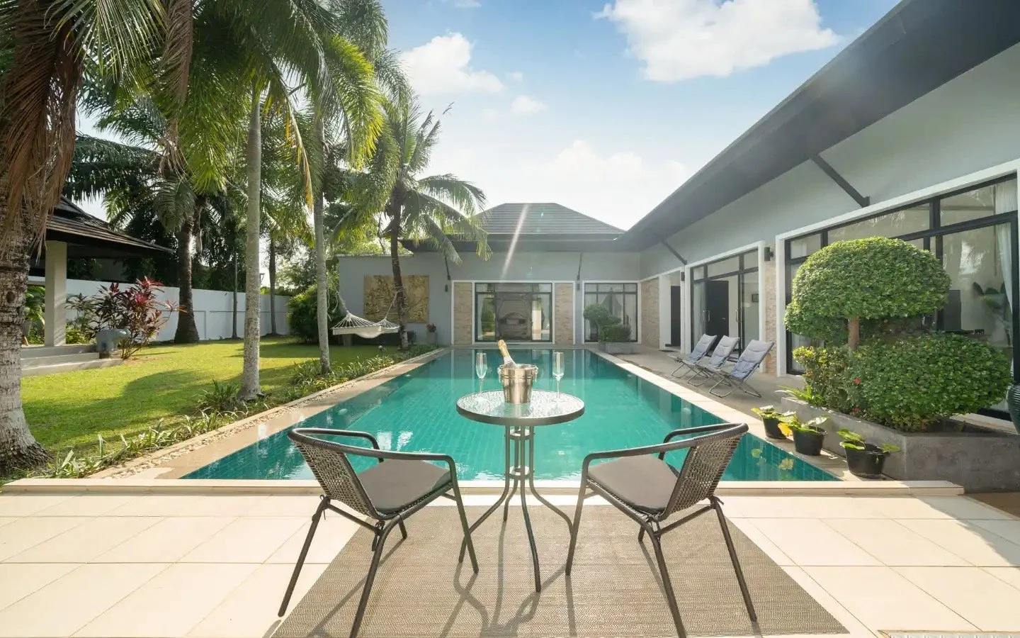 Luxury backyard with swimming pool, patio furniture, lush greenery, and modern house under a sunny sky. Villa Lotus Heaven, Cherng Talay, Phuket