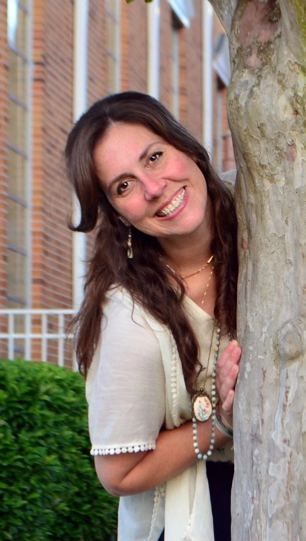 A woman with long brown hair smiling and leaning around a tree. She is wearing a cream-colored top with decorative trim and jewelry, including necklaces and earrings. There is a brick building with large windows and greenery in the background.