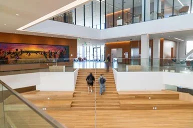 Interior view of a modern building with stairs leading up to a mezzanine level, glass railings, and two people walking up the stairs.